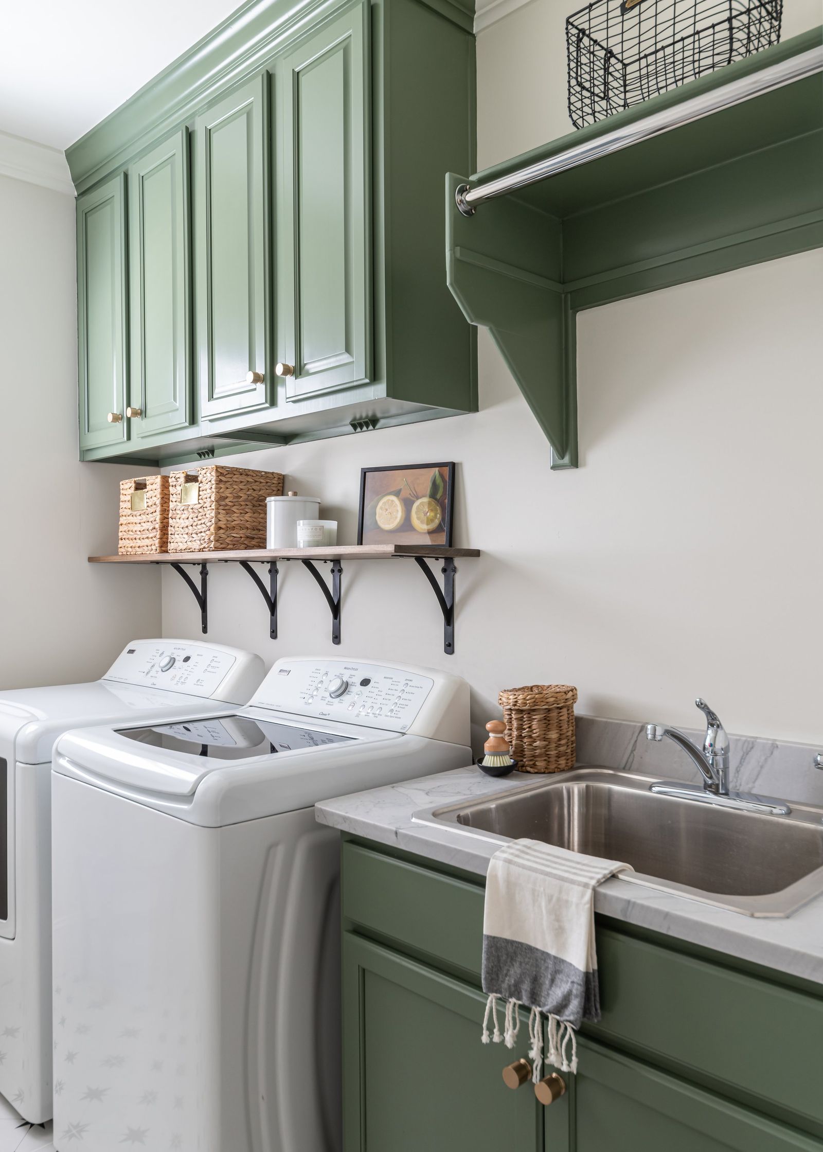 Sage-green laundry cabinets above white machines with an iron-bracket shelf and baskets