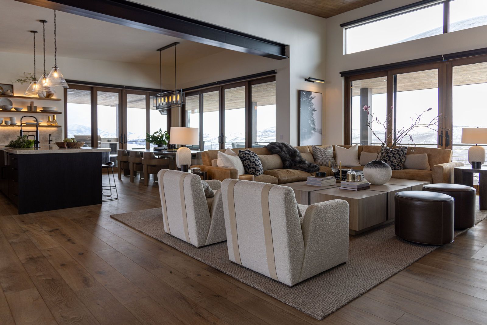 Open great room with cream swivel chairs, a leather sectional and walls of windows onto snowy hills