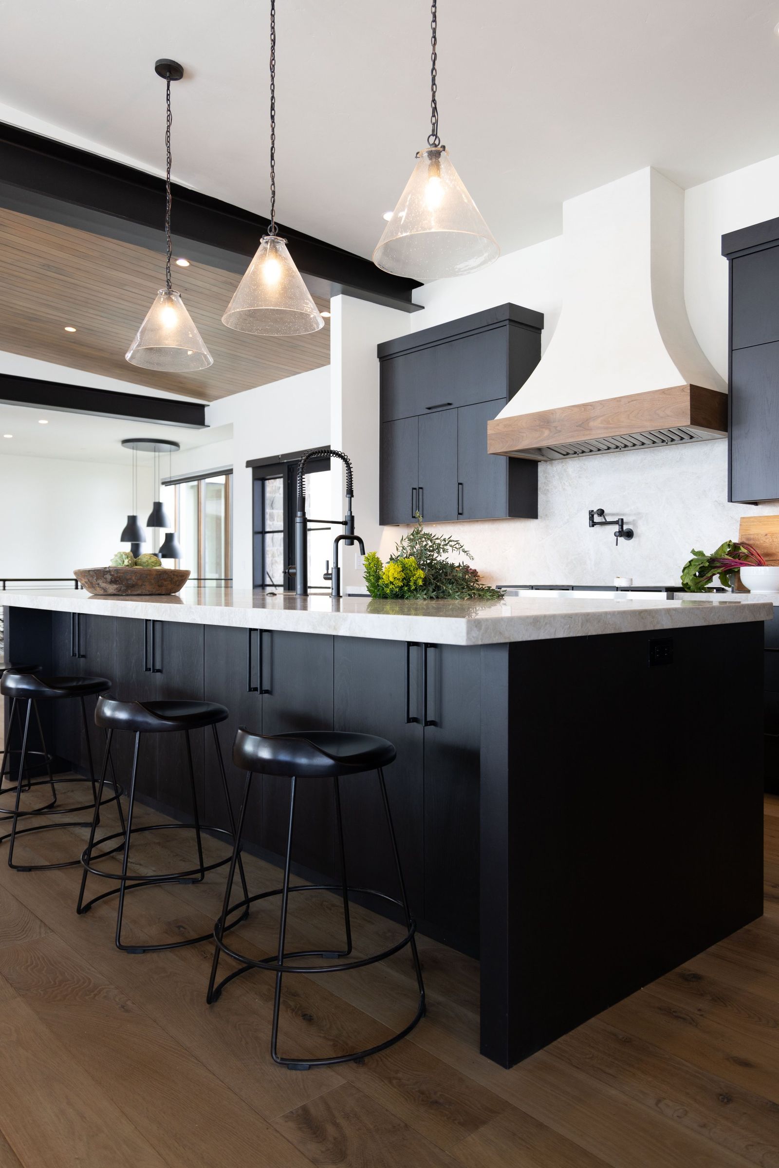 Glass pendants over a charcoal island with black leather stools and a white plaster hood