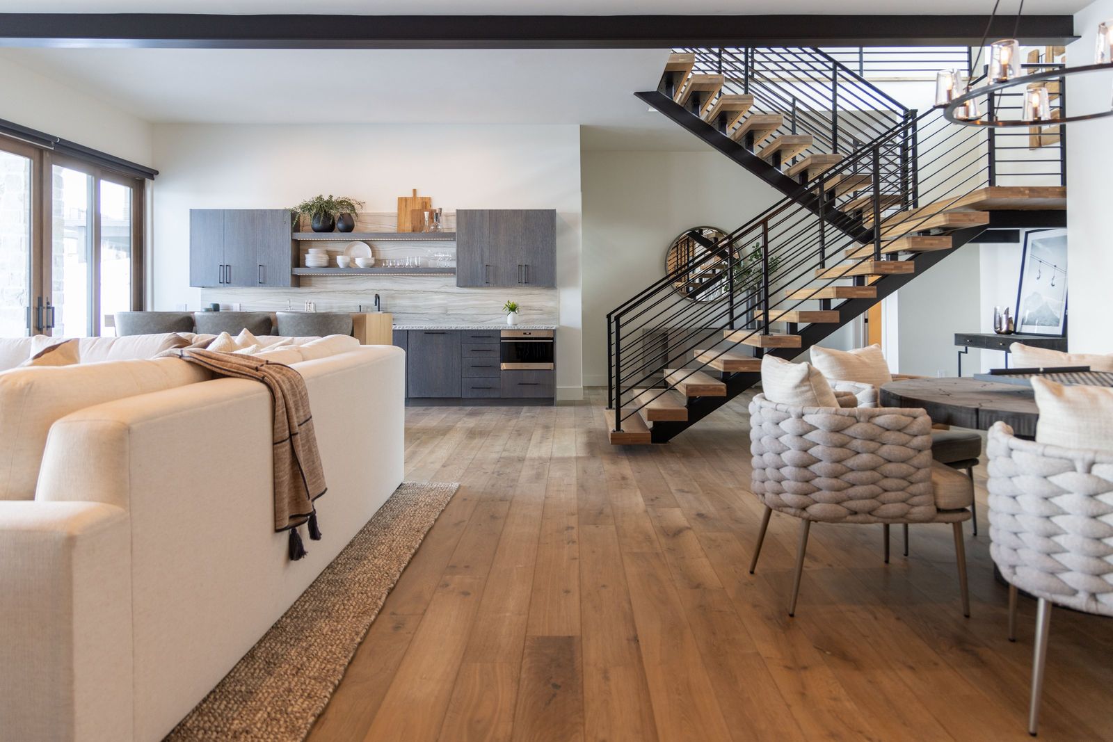 Lower-level kitchenette and floating stair seen past a cream sofa and woven chairs