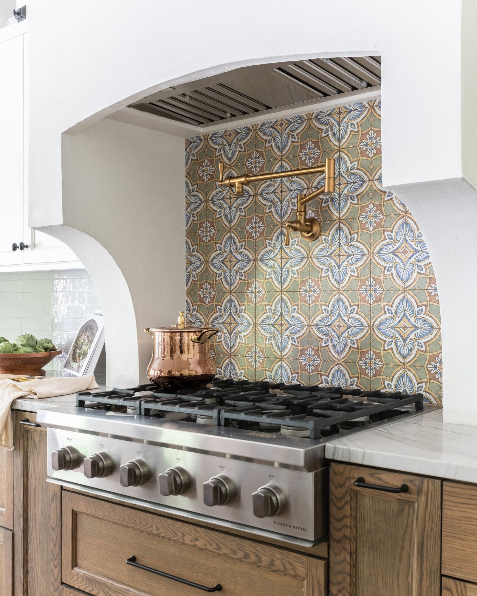 Arched plaster range alcove lined with patterned tile, a brass pot filler and a copper pot