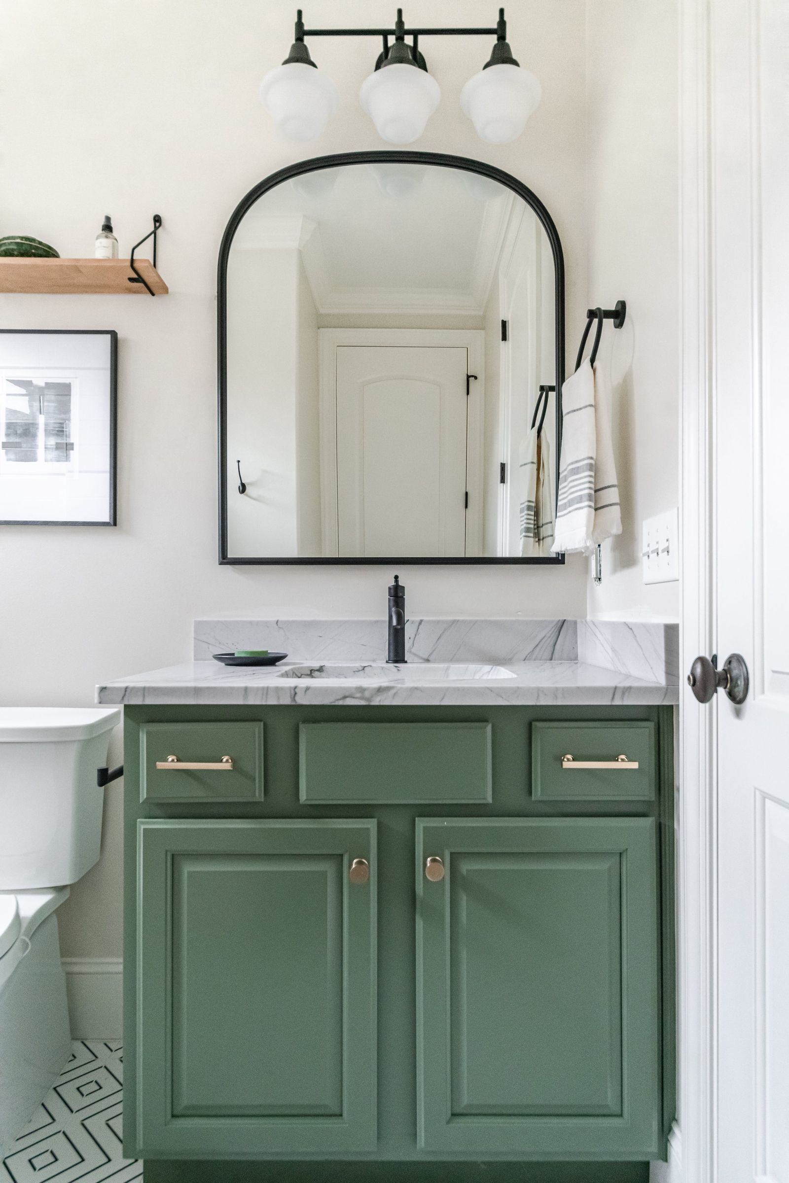 Forest-green vanity with brass pulls under an arched black mirror and globe sconces