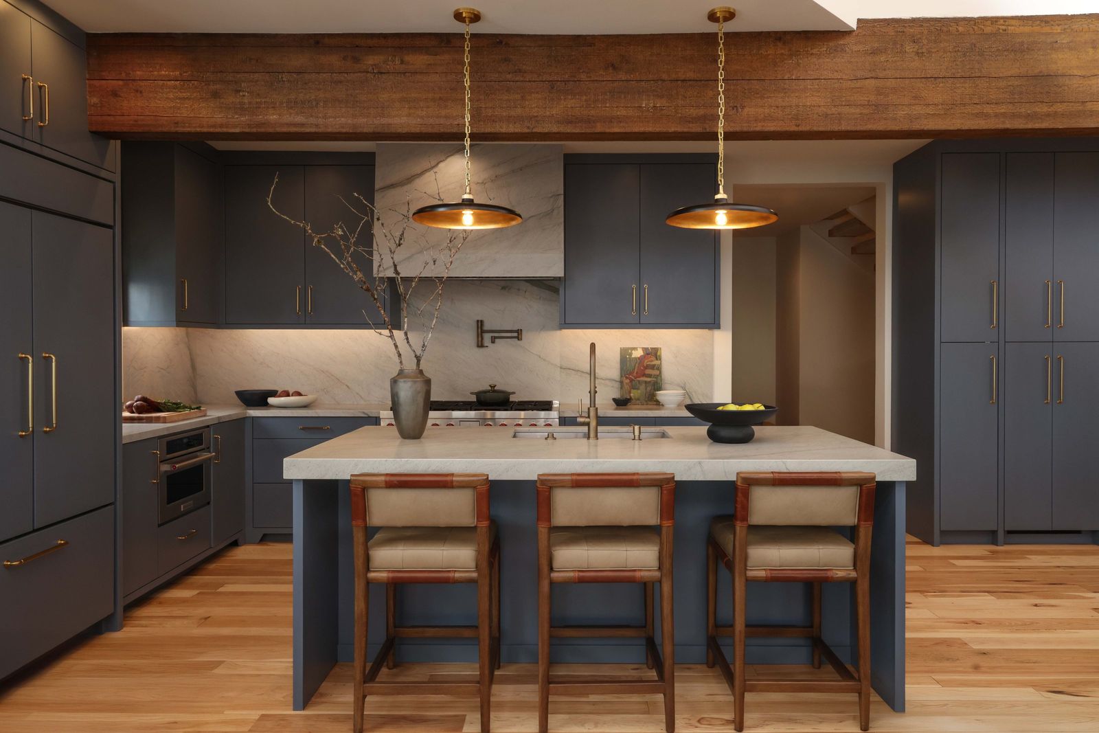 Slate-blue kitchen with a quartzite hood, brass pendants and leather-back stools under a timber beam