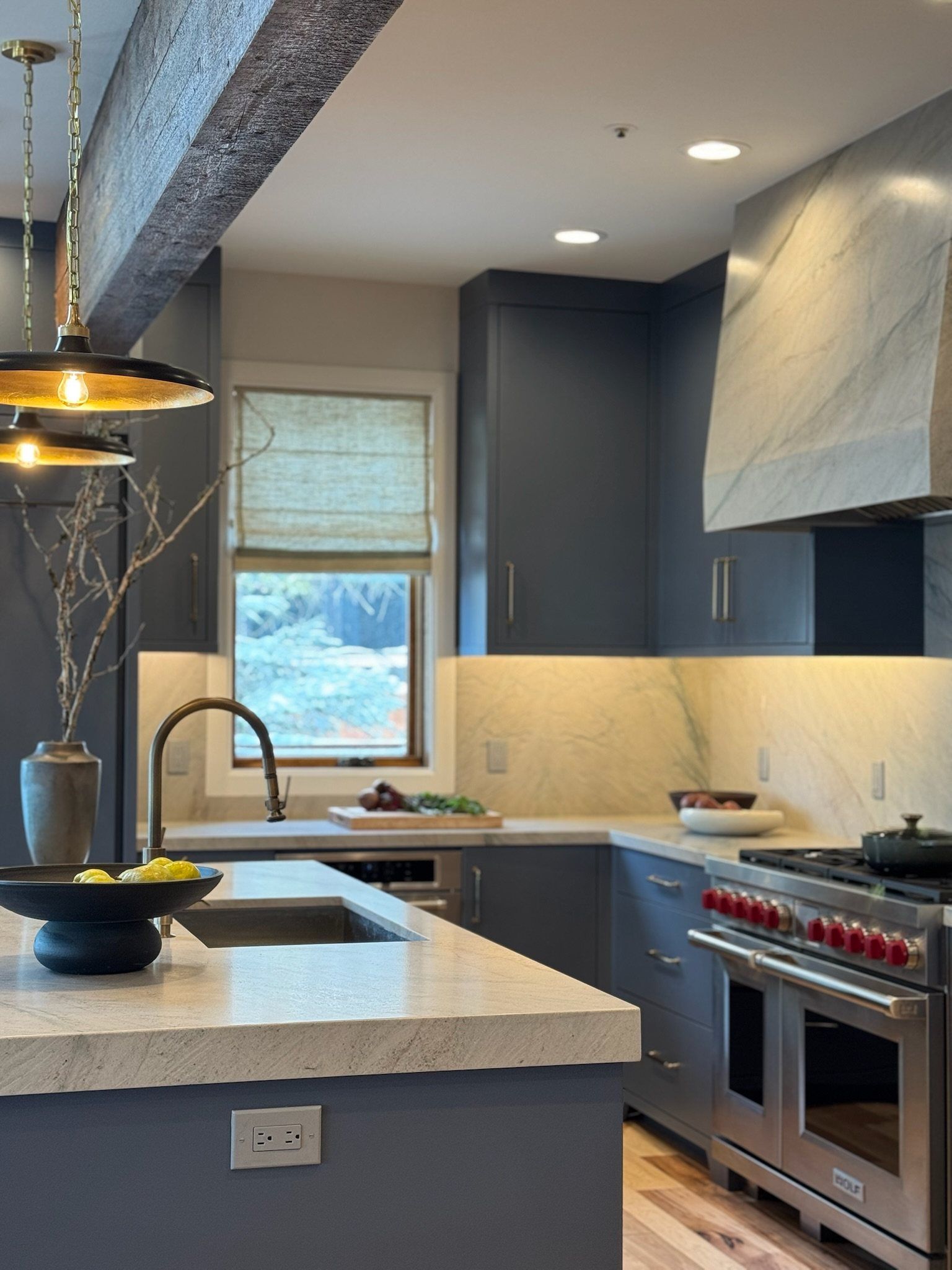 Blue cabinetry, quartzite backsplash and a pro range beside a window with a woven shade
