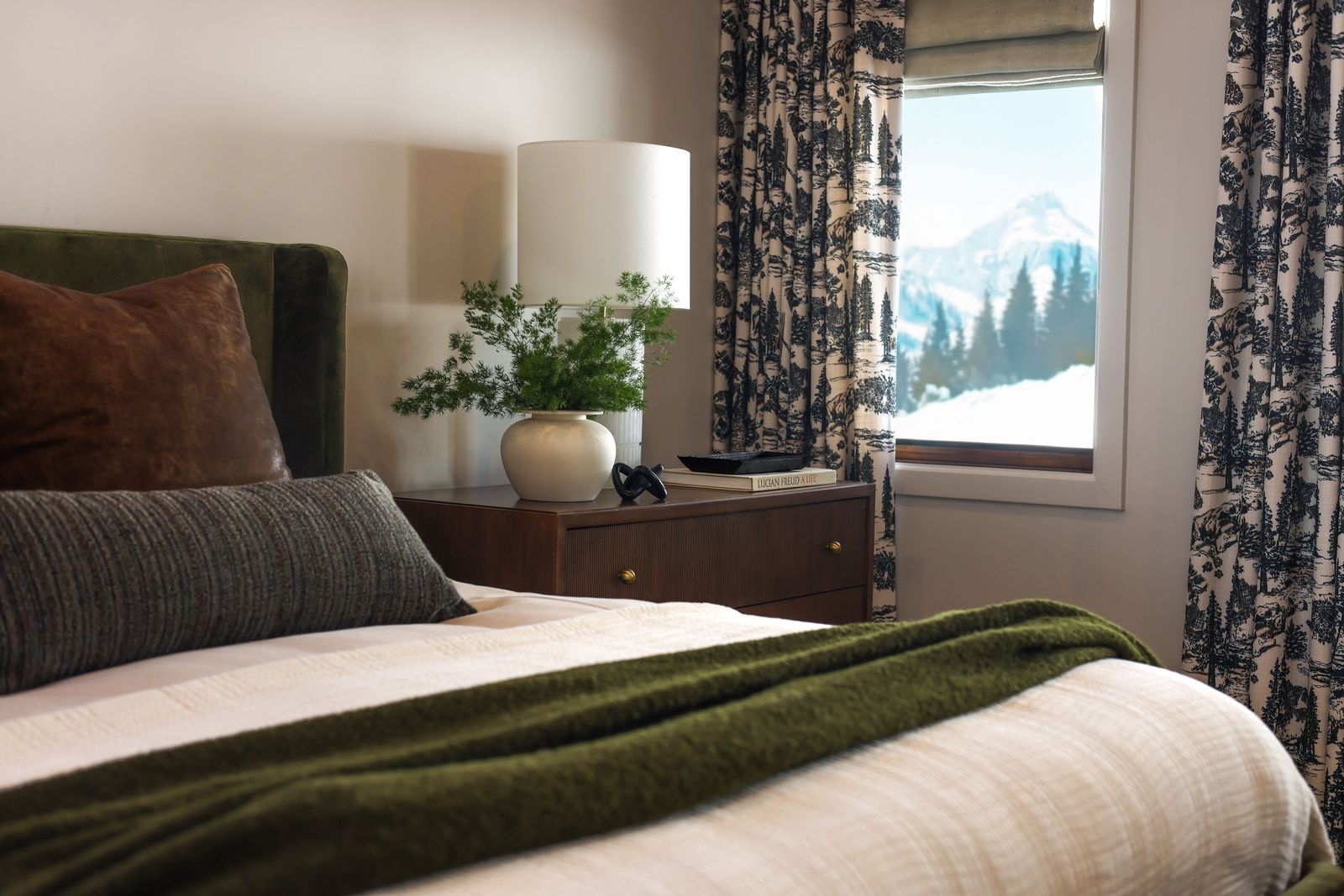 Primary bedroom with a green velvet bed, toile drapery and a window framing snowy peaks