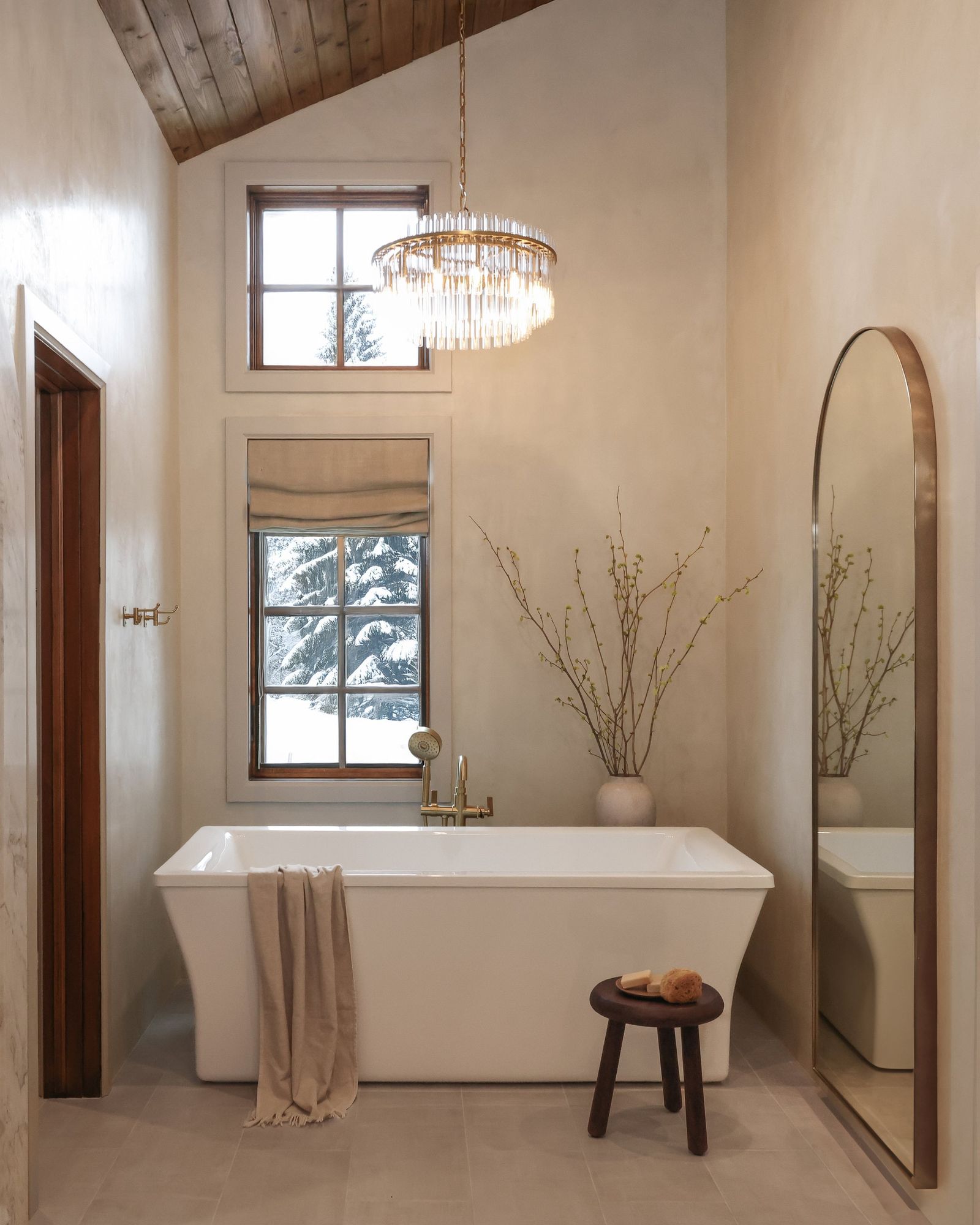 Freestanding tub under a crystal chandelier with a wood stool and tall arched mirror