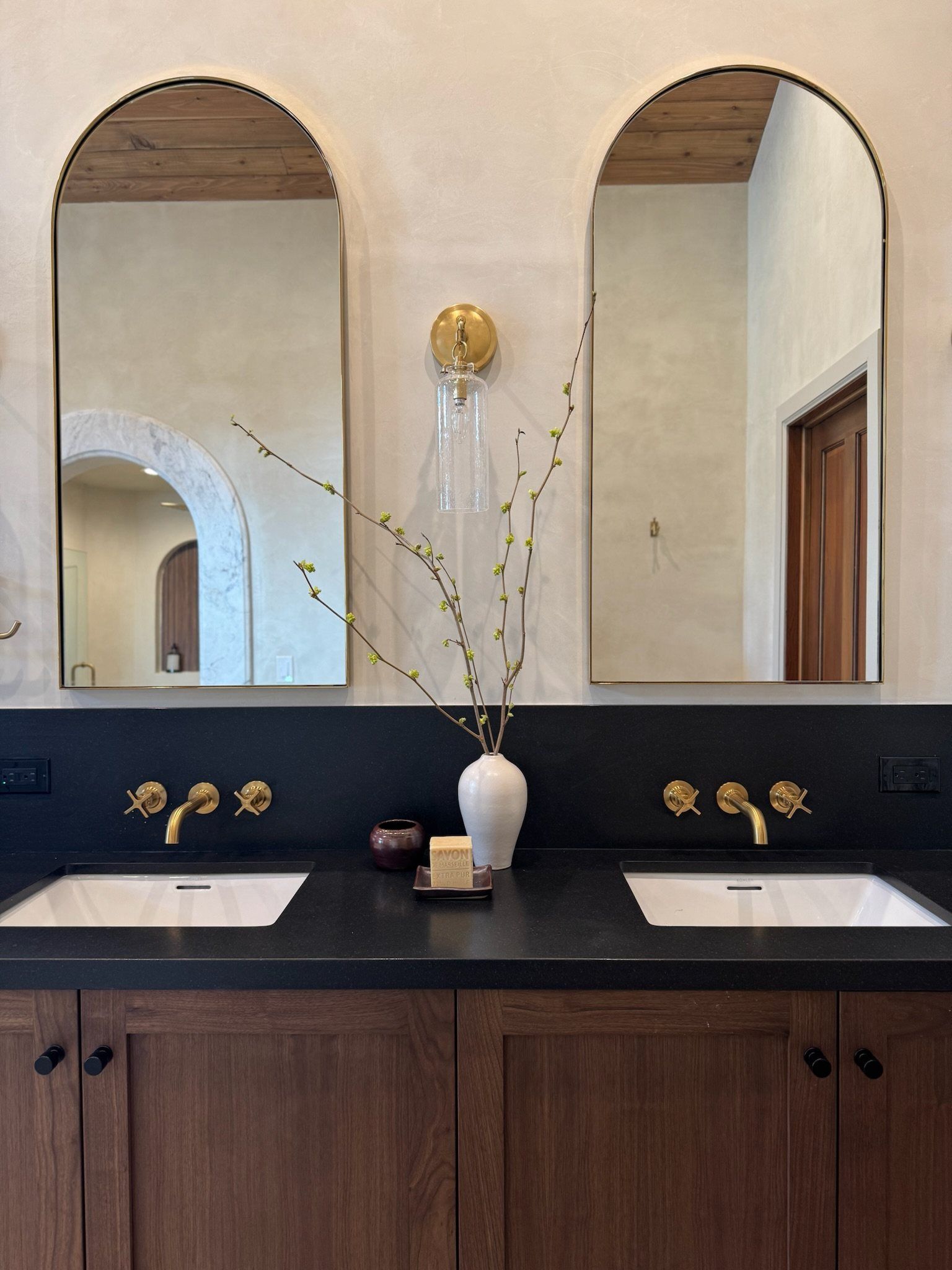 Double vanity with black counters, arched brass mirrors and wall-mount faucets