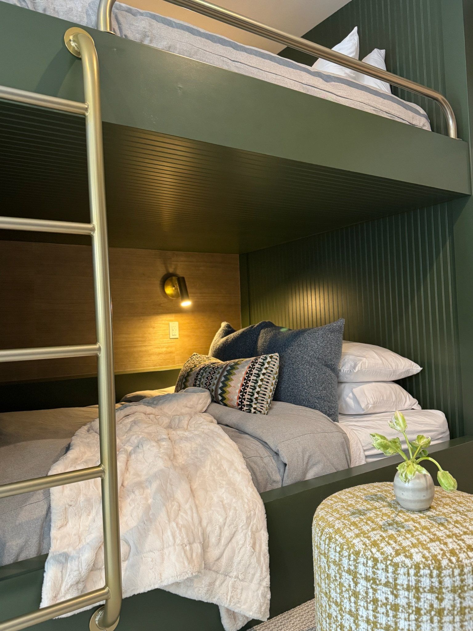 Lower bunk with a warm sconce, navy pillows and a woven pouf beside the brass ladder