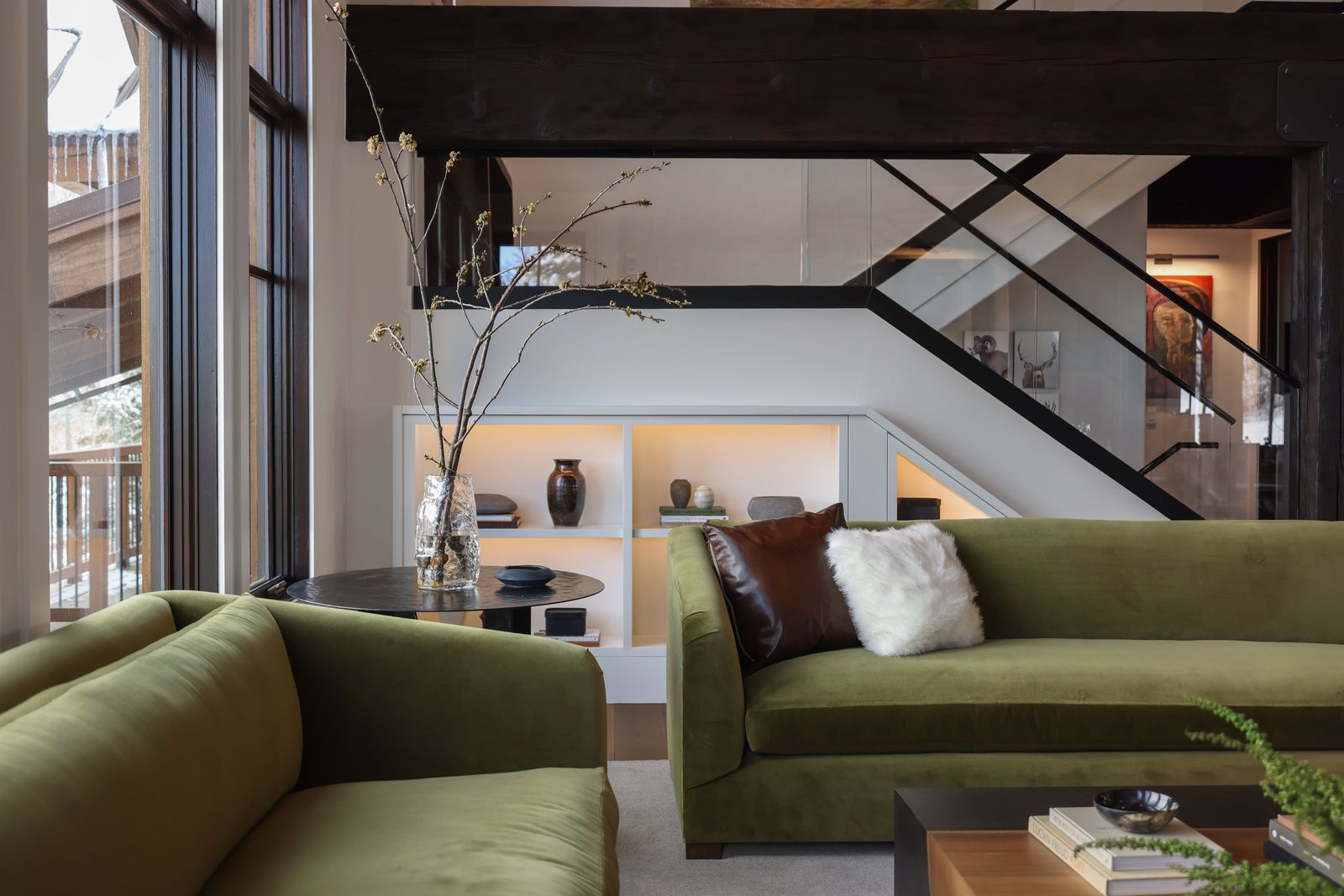 Olive velvet sofas facing a lit built-in beneath a steel and glass staircase