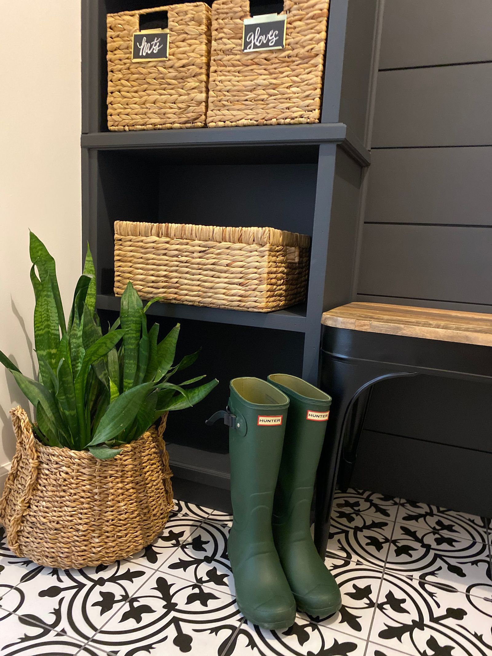 Black cubbies with woven baskets, green rain boots and a snake plant on patterned tile