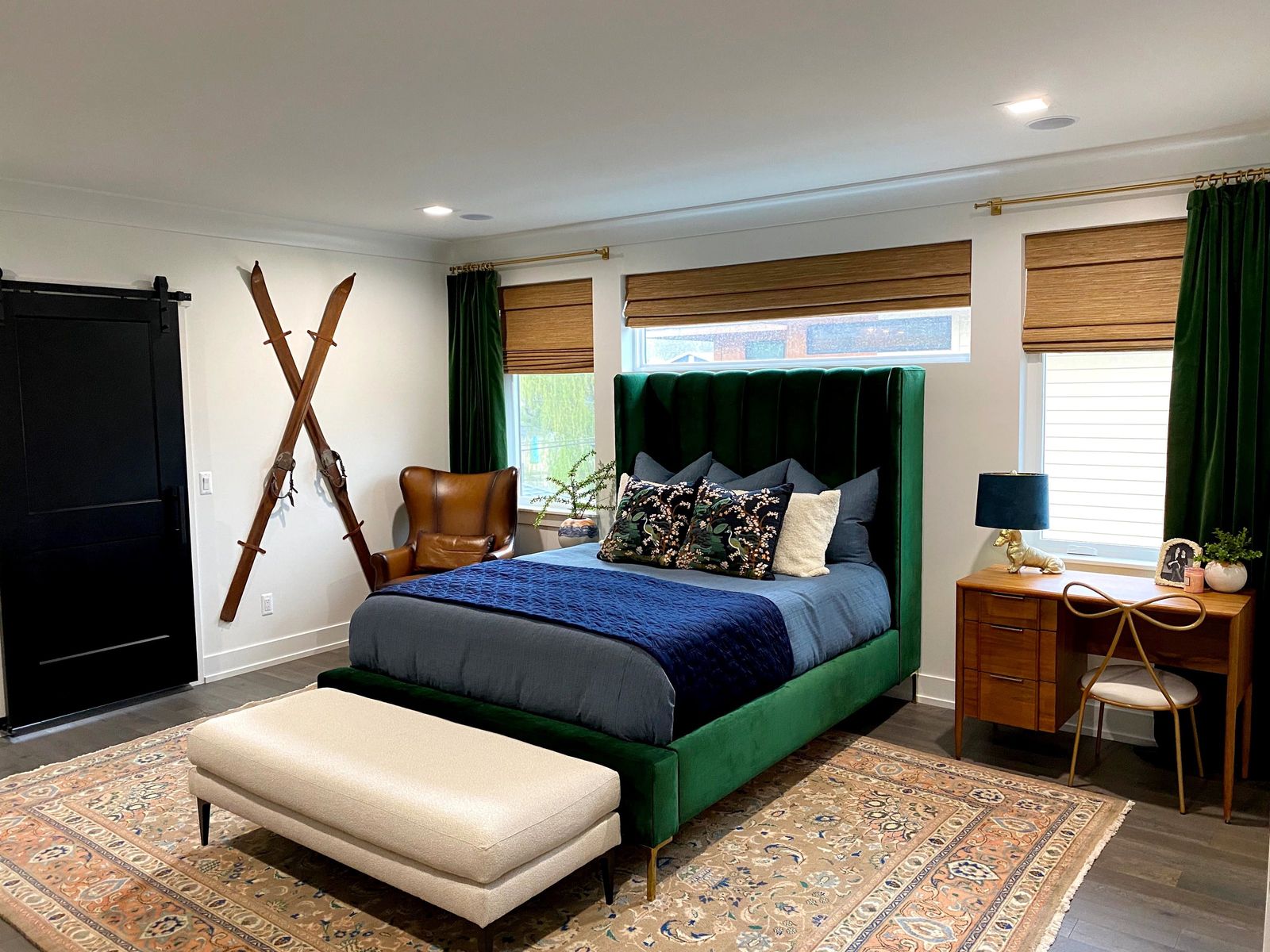 Bedroom with a green velvet channel bed, navy quilt, crossed vintage skis and woven shades