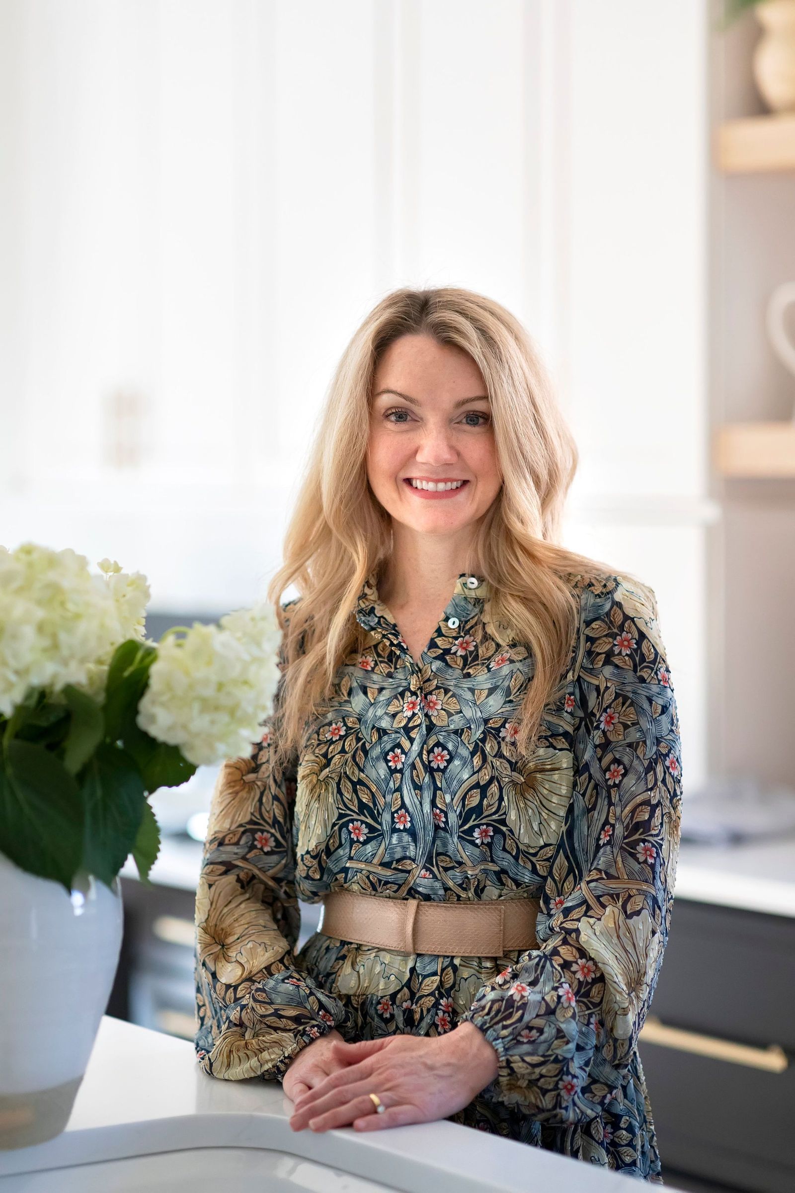 Joni Glass, designer, smiling at a white kitchen counter in a paisley dress